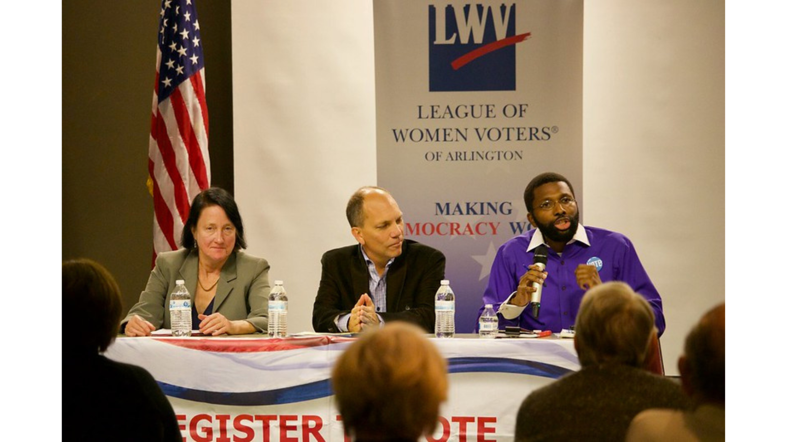 Three candidates are sitting behind a desk at a candidate forum. Behind the candidates is an American flag (left) and League of Women Voters of Arlington vertical banner. The banner has the blue box LWV logo and says: LEAGUE OF WOMEN VOTERS OF ARLINGTON in blue text. Below that is the text: MAKING DEMOCRACY WORK. One of the three candidates is wearing a purple shirt and speaking into a microphone to the audience.