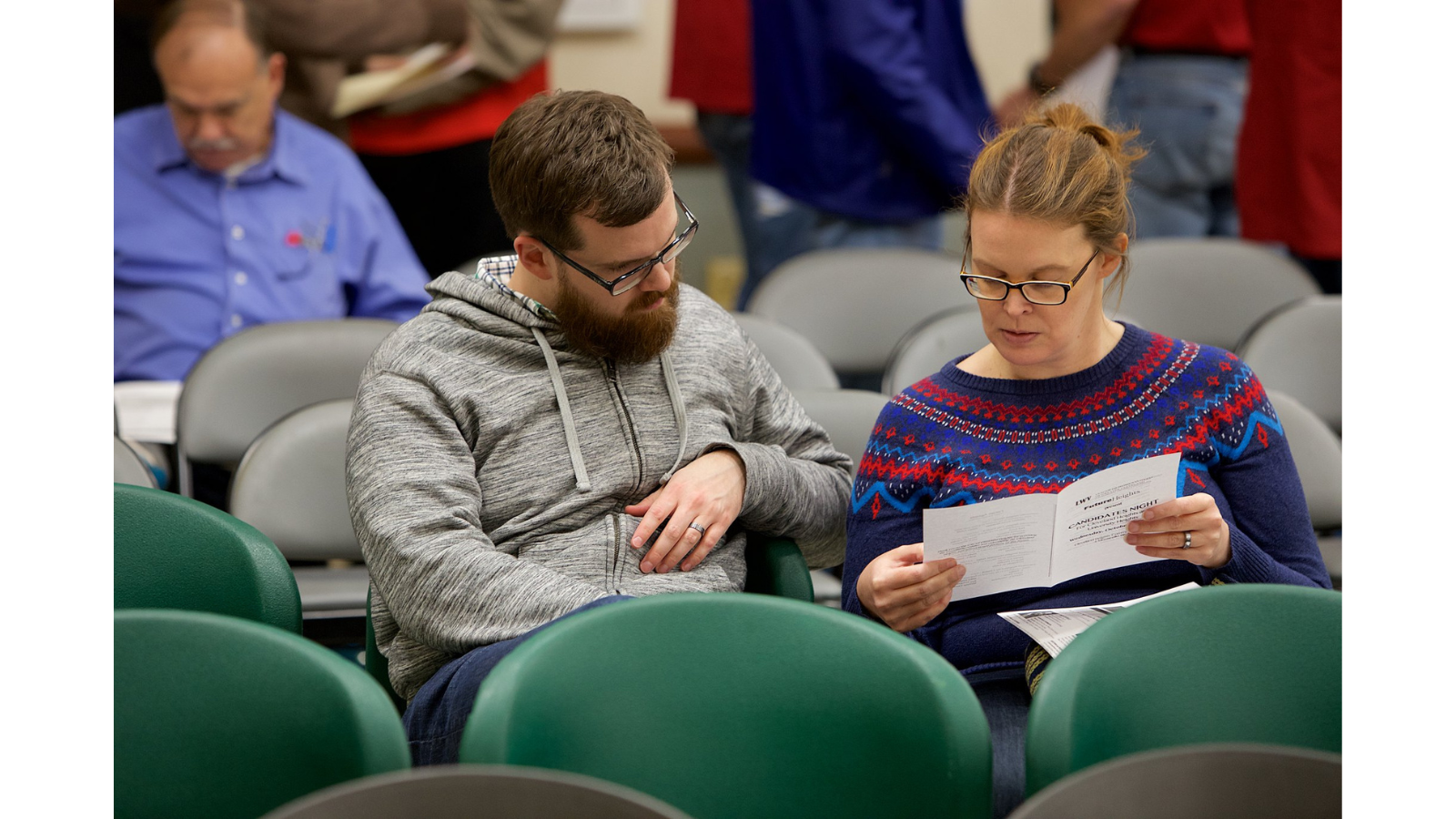 A man and a woman are sitting down at a candidate forum. The woman is holding a League candidate night pamphlet, and the man is looking over at it. The man and woman are reading the pamphlet together.