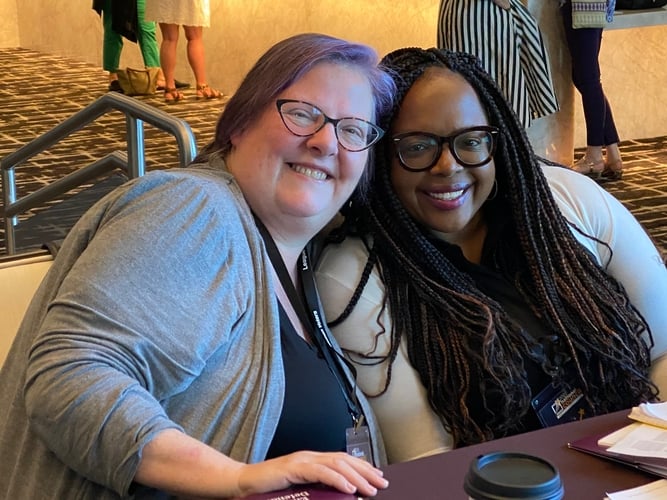 A white woman with purple hair and glasses and a Black woman with braided hair and glasses smile together seated at a table